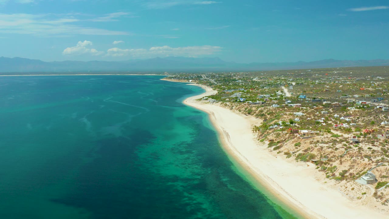 High aerial view over El Sargento La Ventana, Mexico, with turquoise water on the left, a pale sand shoreline curving inland, scattered houses on the right, distant mountains, and no visible people.