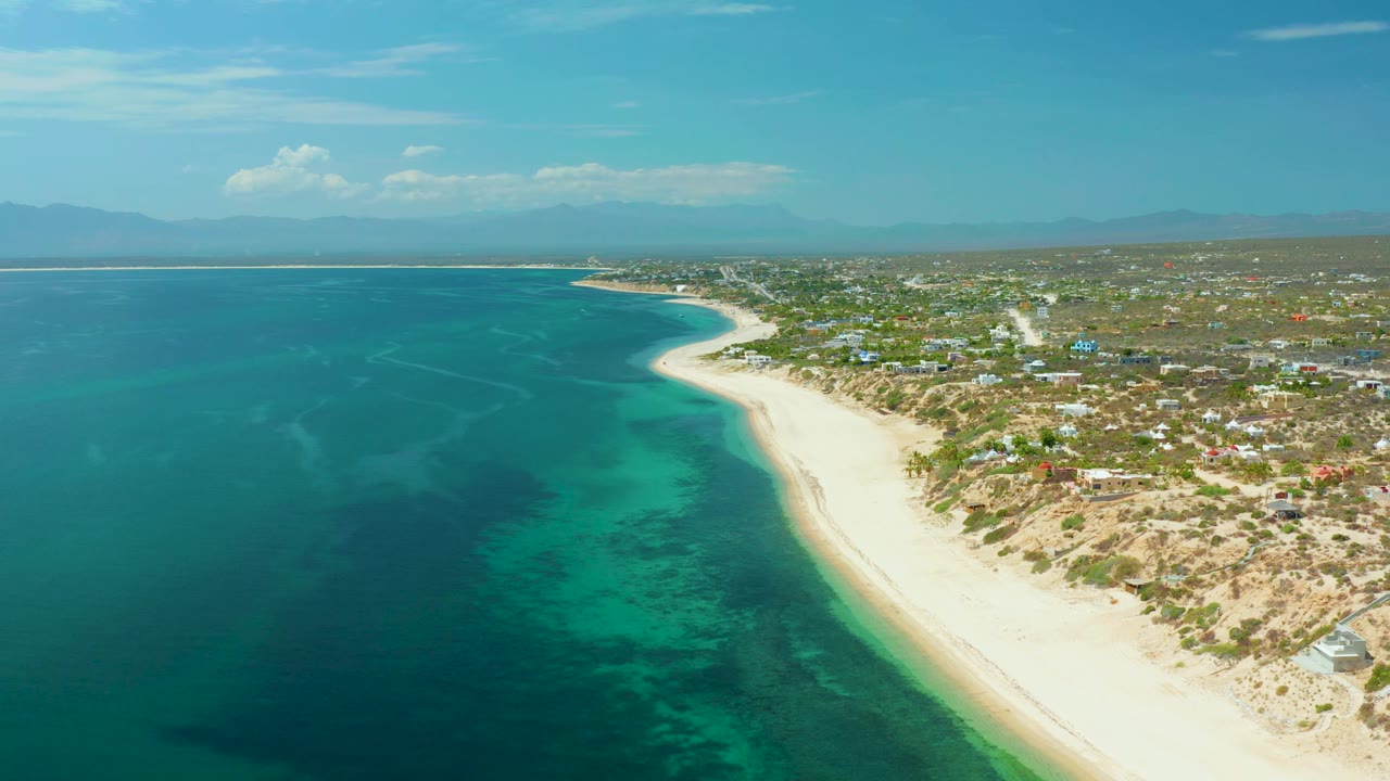 Wide aerial still from El Sargento La Ventana, Mexico, showing darker patches beneath the ocean surface, a bright sand strip, low coastal buildings, distant mountains, and no visible people.