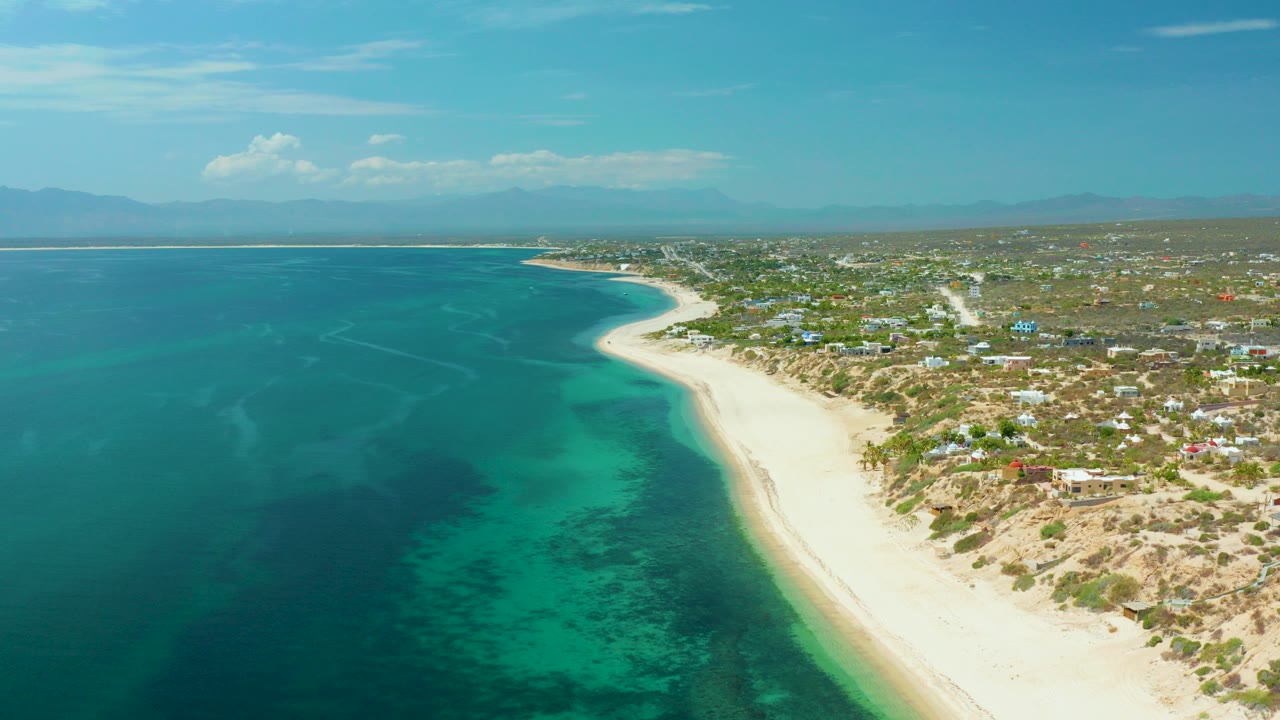 High aerial frame over El Sargento La Ventana, Mexico, with turquoise water on the left, a bright sand beach curving along the coast, low buildings inland, hazy mountains beyond, and no visible people.