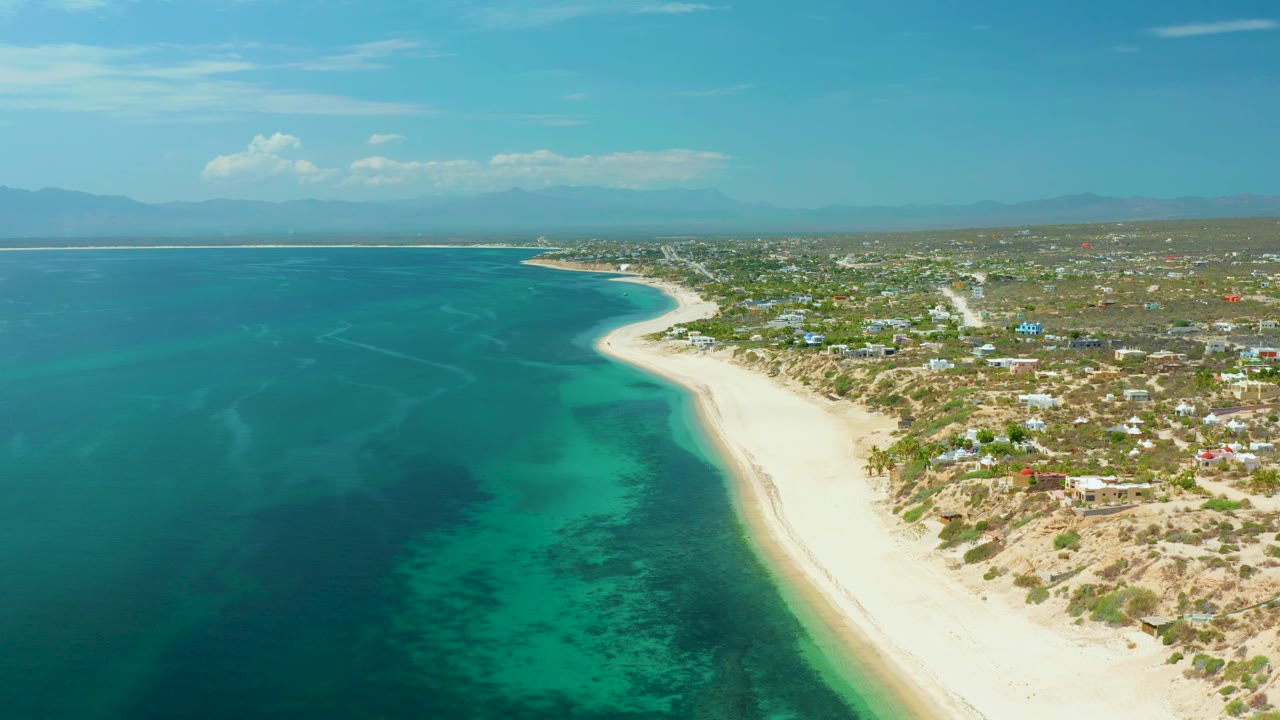 High coastal frame from El Sargento La Ventana, Mexico, with the sea filling the left side, a sweeping sand strip through the center, low inland structures to the right, soft clouds overhead, and no visible people.