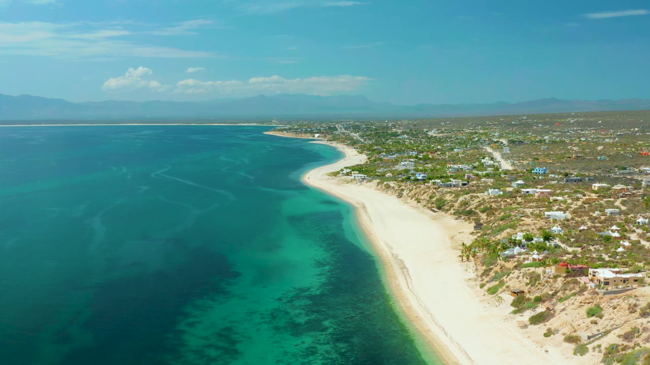 Wide drone view of turquoise shallows, beach curve, dunes, and houses along the coast.