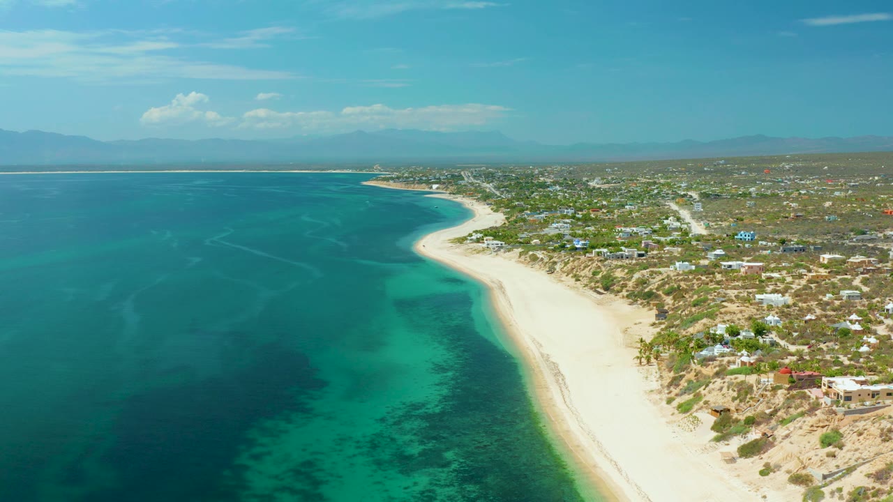 Aerial frame over El Sargento La Ventana, Mexico showing a longer stretch of green shallows against pale sand, low homes and vegetation to the right, hazy mountains in the distance, and no visible people.