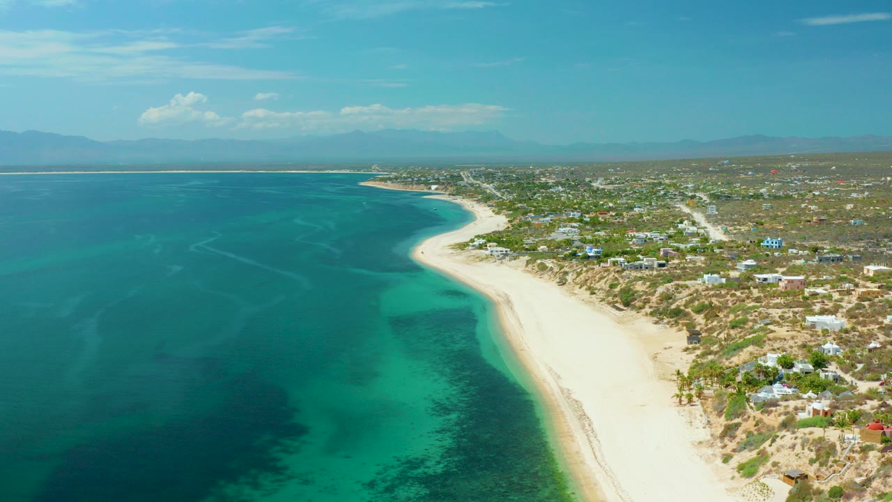 Aerial view over El Sargento La Ventana, Mexico, with turquoise ocean on the left, a pale beach curving into the distance, low coastal buildings inland, distant mountains, and no visible people.