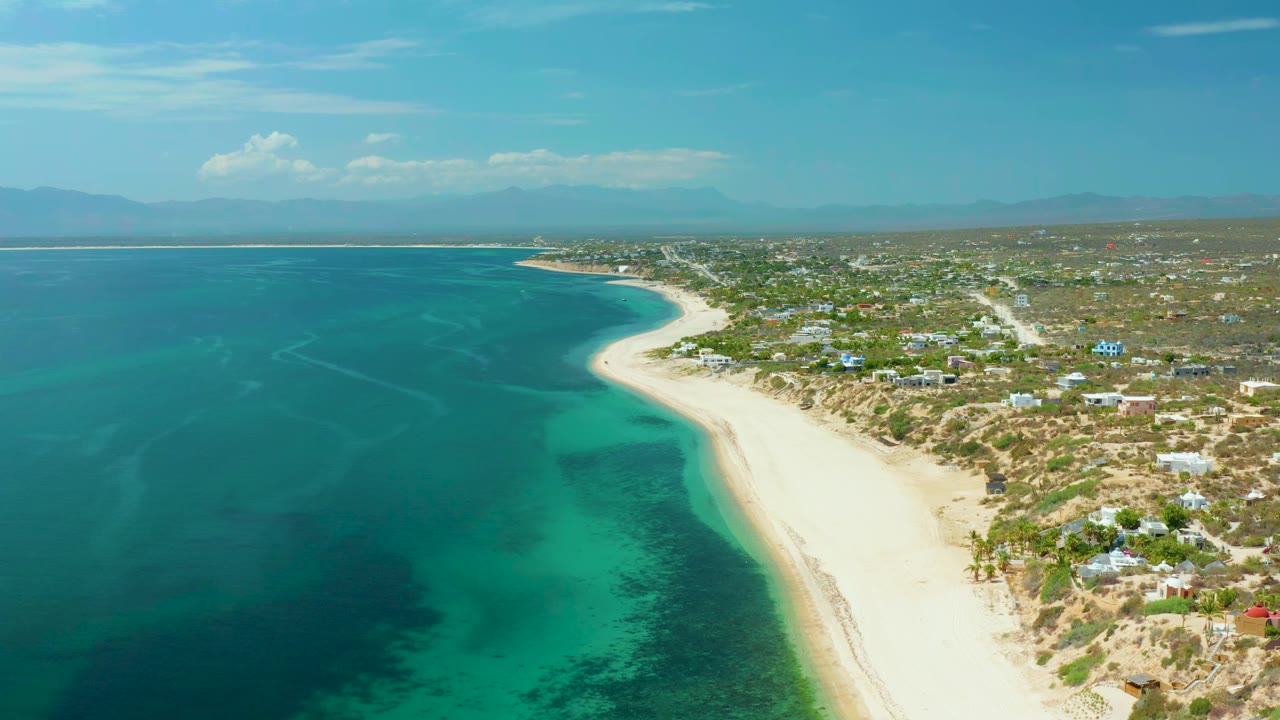 High coastal view with blue ocean, pale beach, dunes, and scattered buildings.