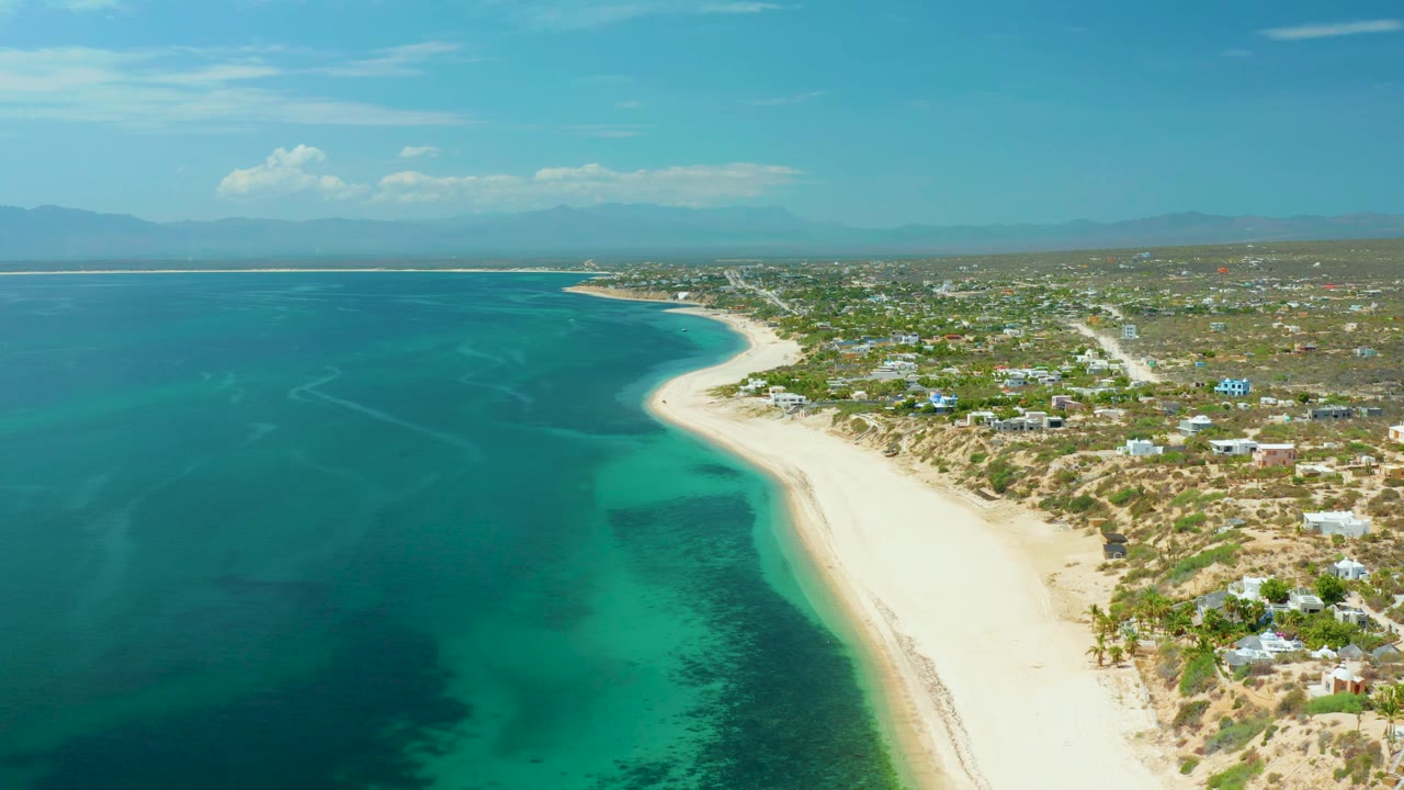 Drone view looking along a sandy coast with turquoise water and low buildings inland.