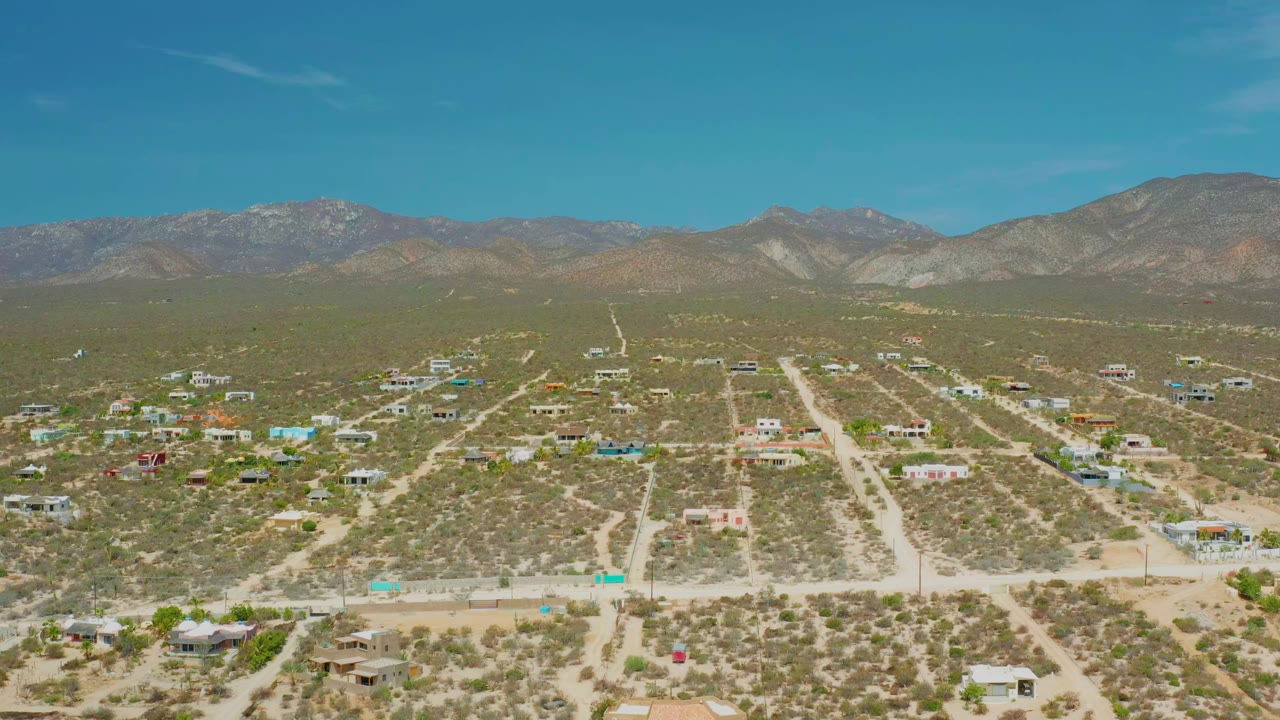 Opening aerial frame over El Sargento La Ventana, Mexico, showing scattered desert homes, pale dirt roads, a mountain horizon, blue sky, and no visible people.