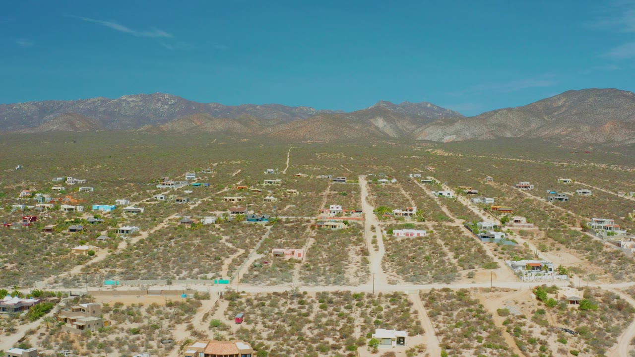 Opening aerial frame over El Sargento La Ventana, Mexico, showing pale dirt roads through scrubland, scattered homes, distant mountains, blue sky, and no visible people.