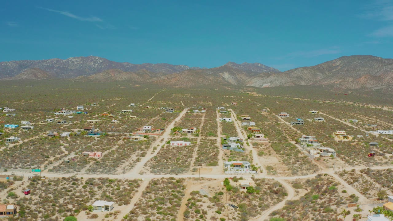 Opening aerial frame over El Sargento La Ventana, Mexico, showing dusty roads, sparse homes, scrub terrain, distant mountains, blue sky, and no visible people.