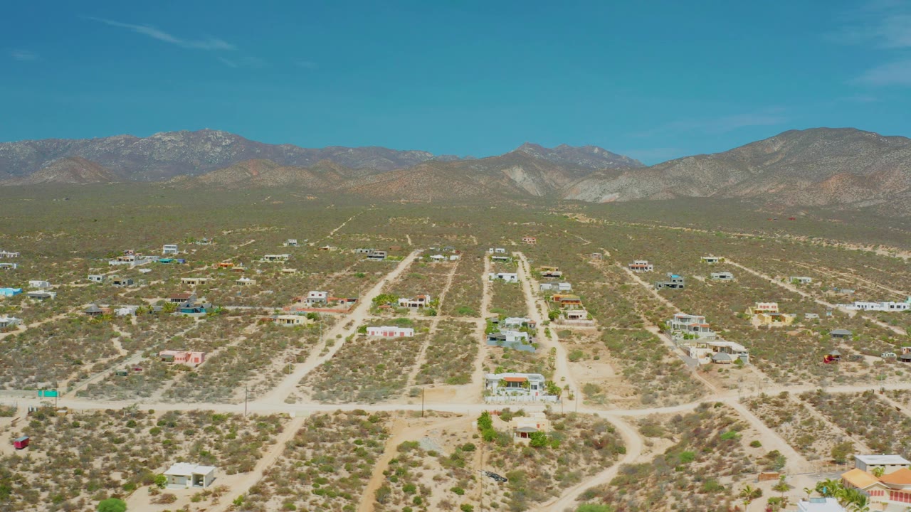 Third frame in El Sargento La Ventana, Mexico, looking across pale dirt roads, low houses, green-brown scrub, a continuous mountain ridge, and no visible people.