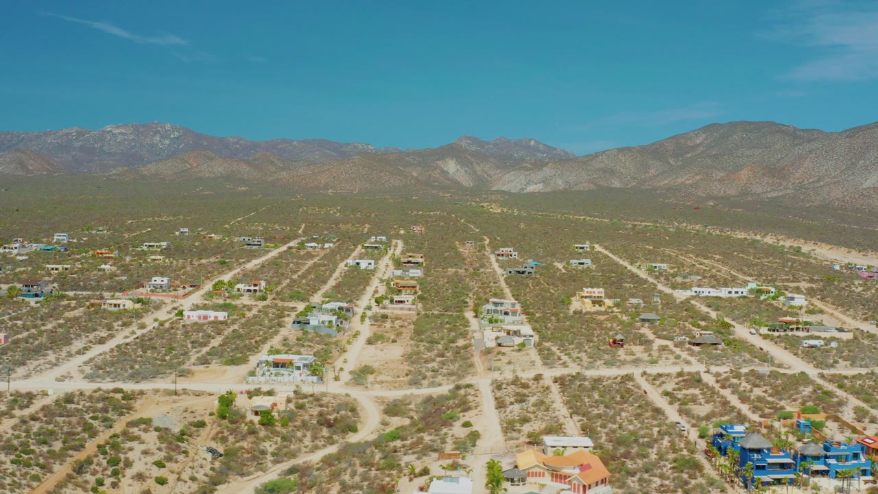 Opening aerial frame over El Sargento La Ventana, Mexico, with pale dirt roads, scattered desert homes, low scrub, distant mountains, blue sky, and no visible people.