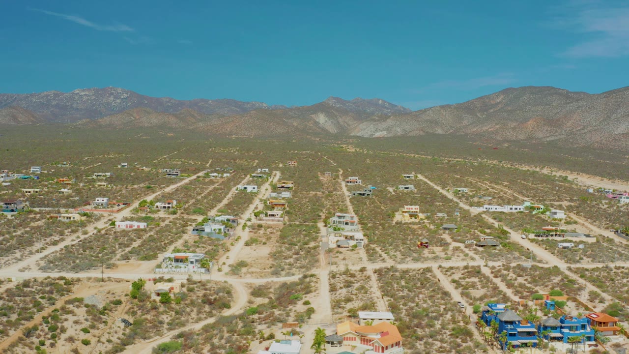Aerial frame over sparse homes and sandy road tracks in El Sargento La Ventana, Mexico, with the mountain ridge filling the background and no visible people.