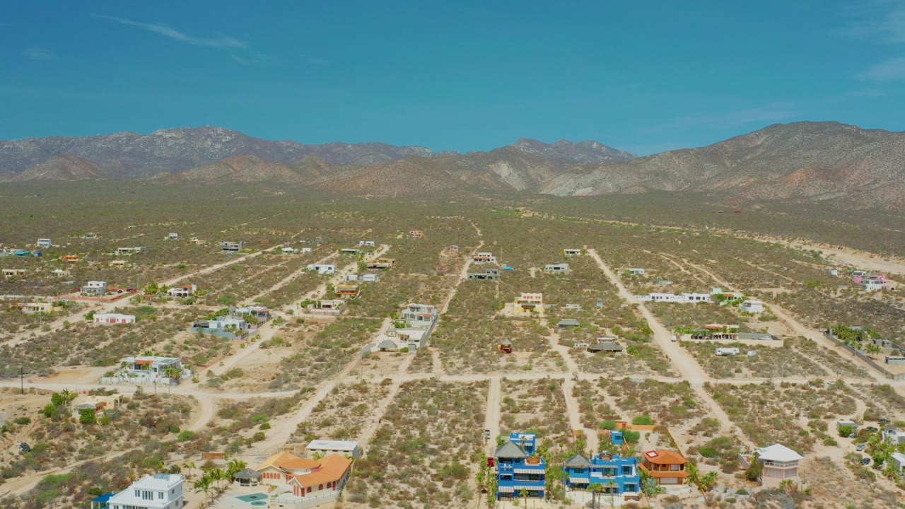 Wide aerial frame over El Sargento La Ventana, Mexico, with scattered homes, pale dirt roads, scrubland, distant mountains, clear blue sky, and no visible people.
