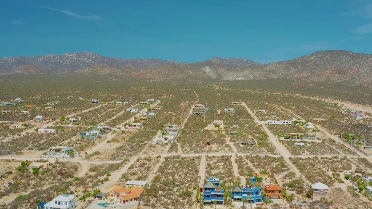 Aerial village grid with sandy roads, scattered houses, scrub vegetation, and mountains.
