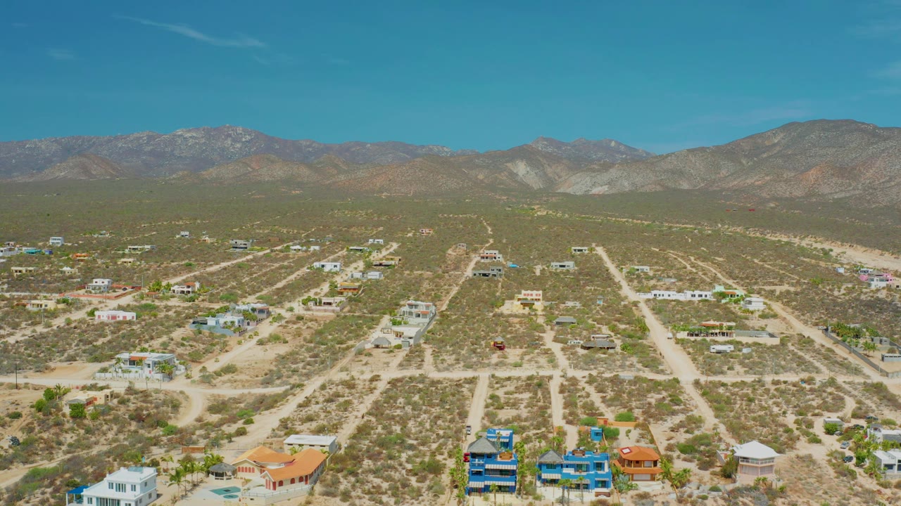 Drone-height frame over El Sargento La Ventana, Mexico, with desert homes and light-colored roads spread across the valley floor below a broad mountain range and no visible people.