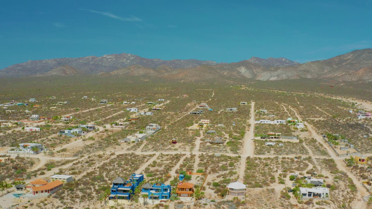 Opening aerial frame over El Sargento La Ventana, Mexico, with dirt roads, scattered homes, desert shrubs, mountain ridges, blue sky, and no visible people.