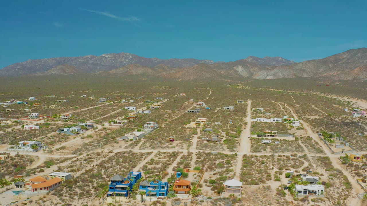 Aerial frame over El Sargento La Ventana, Mexico, with the central dirt road more prominent, low homes spread across desert lots, mountains behind, and no visible people.