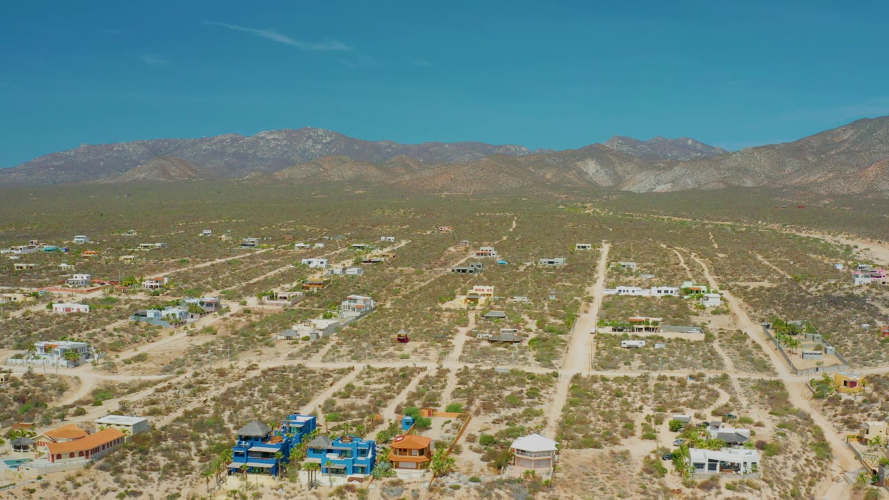 Wide drone frame over El Sargento La Ventana, Mexico, showing sparse desert development, colorful houses near the lower edge, pale roads, mountain ridges, and no visible people.