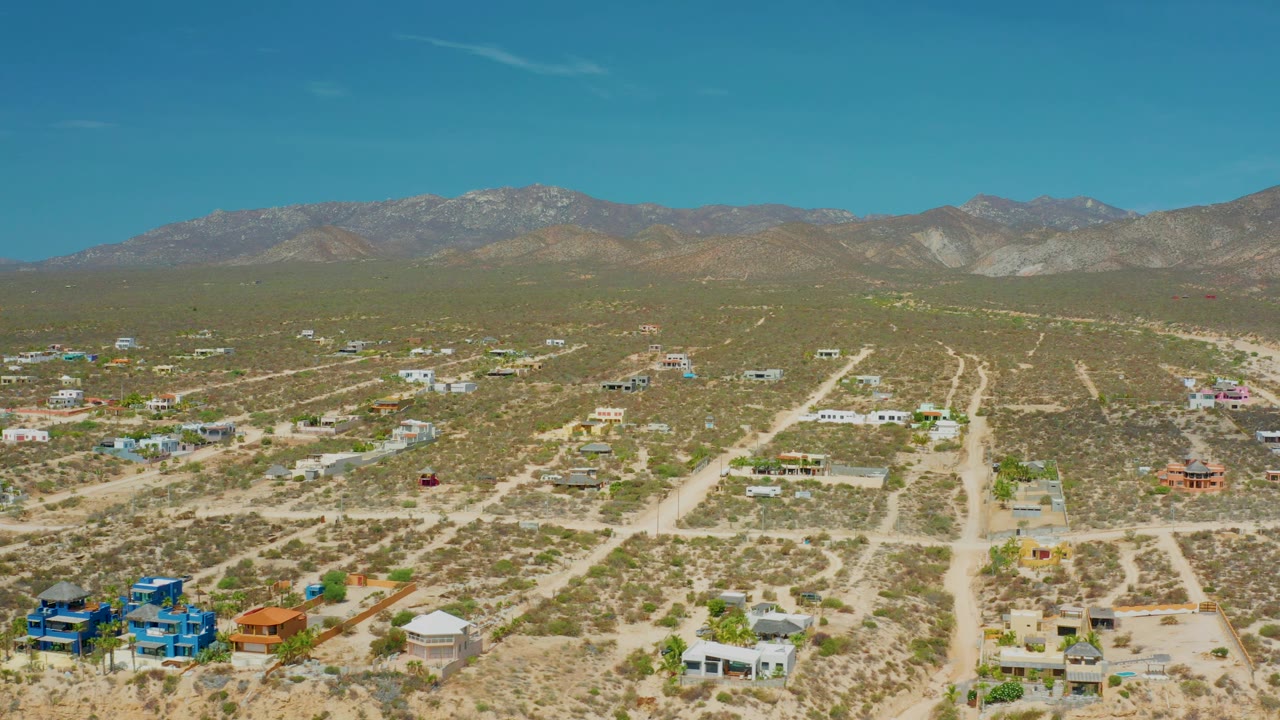 Aerial frame over El Sargento La Ventana, Mexico, with a bright blue building near the lower edge, sandy roads, sparse buildings, distant mountains, and no visible people.