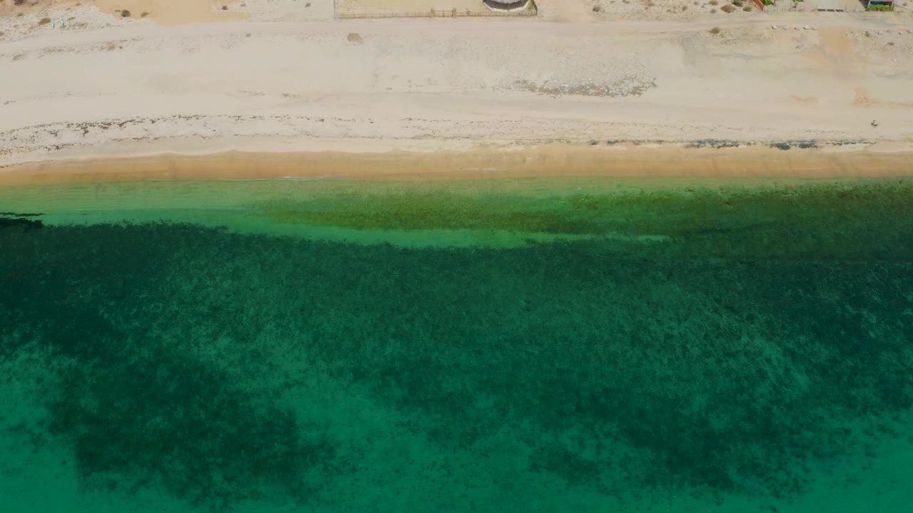 Overhead view of pale beach and clear green water at El Sargento La Ventana, Mexico, with darker underwater patches and no visible people.