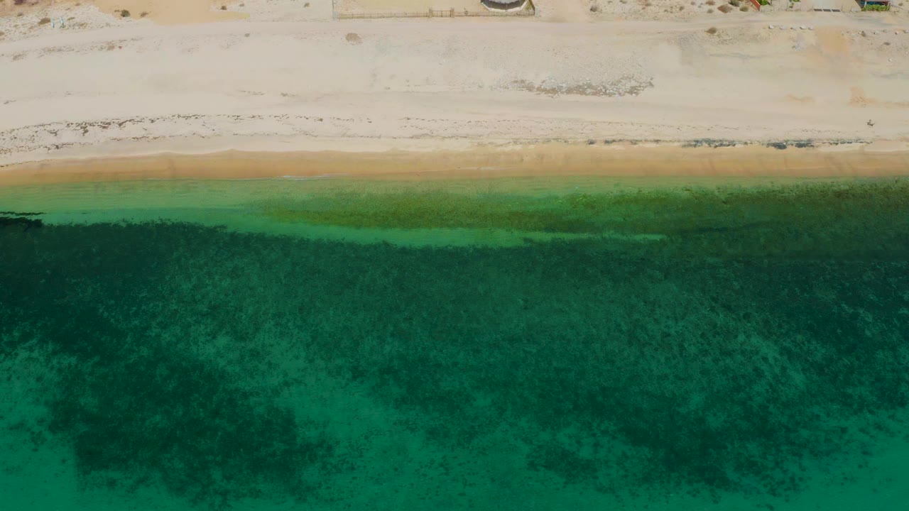 Aerial frame of the beach edge at El Sargento La Ventana, Mexico, where pale sand meets shallow green water with no visible people.