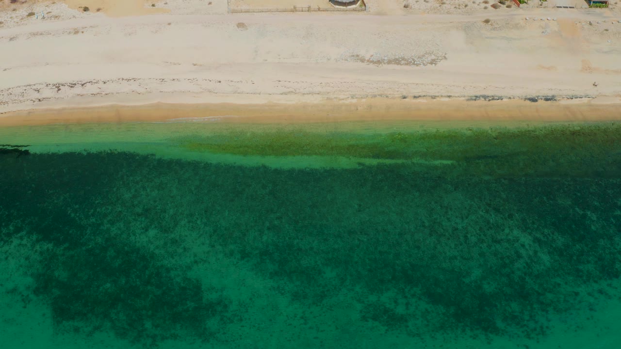 Top-down coastal view at El Sargento La Ventana, Mexico, with clear green ocean over darker seabed areas and the sandy shore across the top, no visible people.
