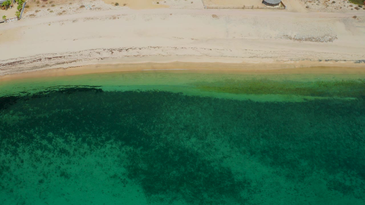 Top-down aerial view of pale sand and clear green ocean at El Sargento La Ventana, Mexico, with darker underwater patches near shore and no visible people.