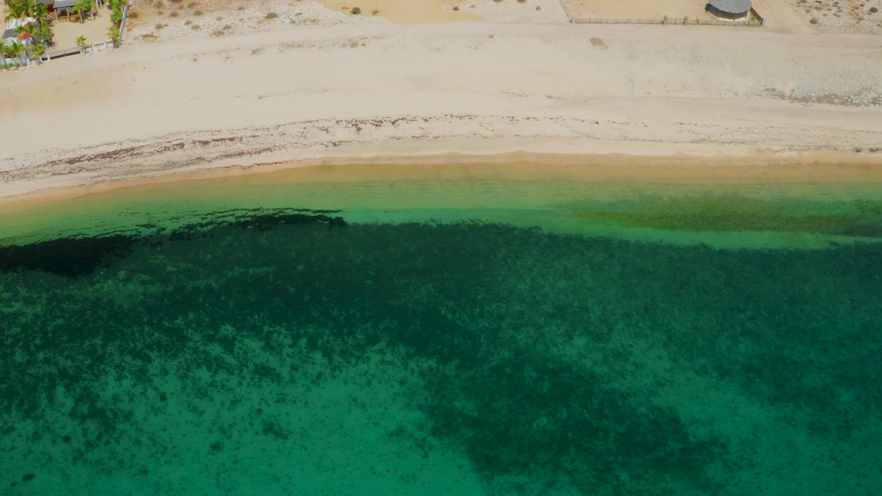 Top-down view at El Sargento La Ventana with a larger dark underwater patch near the left side of the green shallows and empty pale sand above, with no visible people.