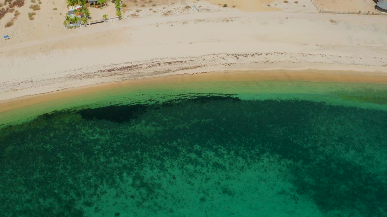 High aerial view over El Sargento La Ventana with pale beach sand above clear green water, dark underwater patches near the surf line, and no visible people.