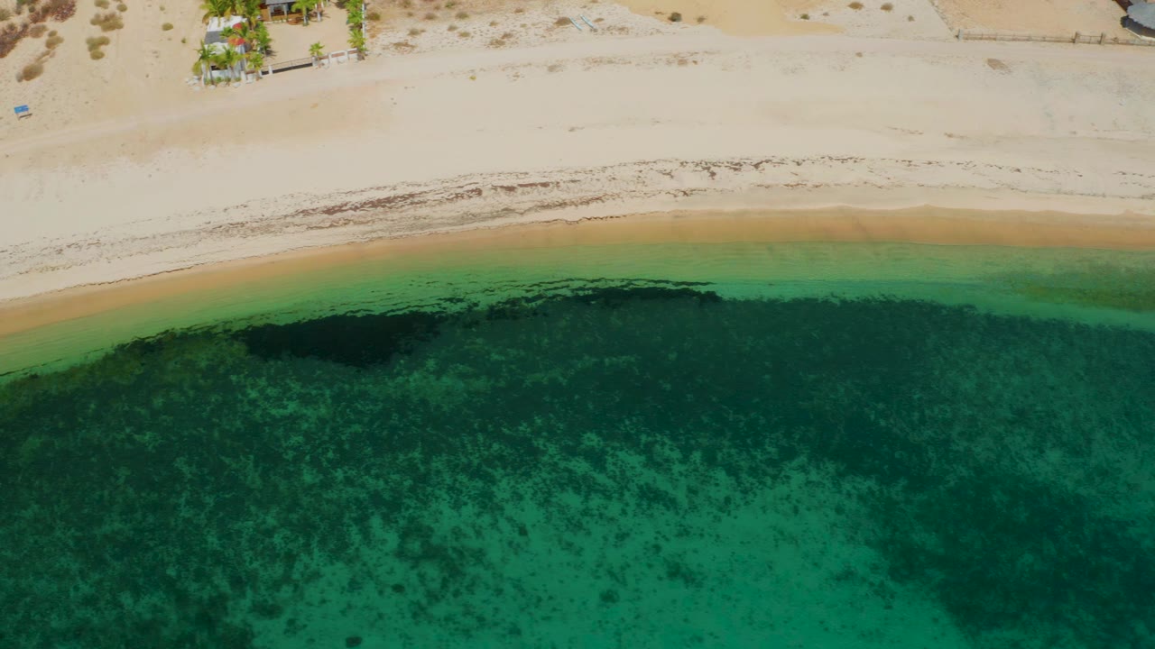 Drone-like top-down frame over El Sargento La Ventana showing a narrow sandy beach, green shallows, one larger dark patch under the water, and no visible people.