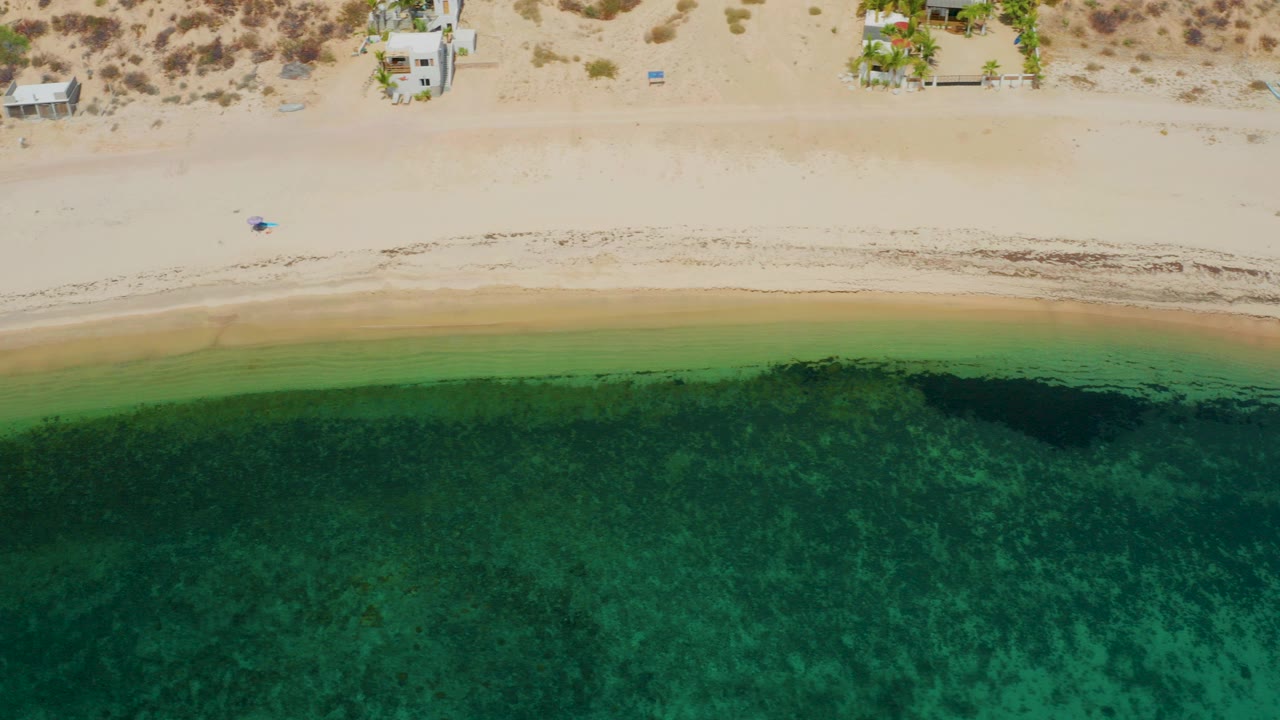 At 0:00, a top-down aerial view in El Sargento La Ventana, Mexico shows pale sand, green shallow water, darker algae near the surf line, scattered beachfront buildings, and no visible people.