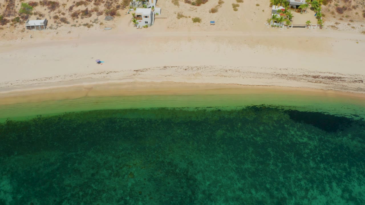 At 0:02, El Sargento La Ventana appears from above with green shallows filling most of the frame, a pale sandy strip, darker algae near the waterline, beach houses, and no visible people.