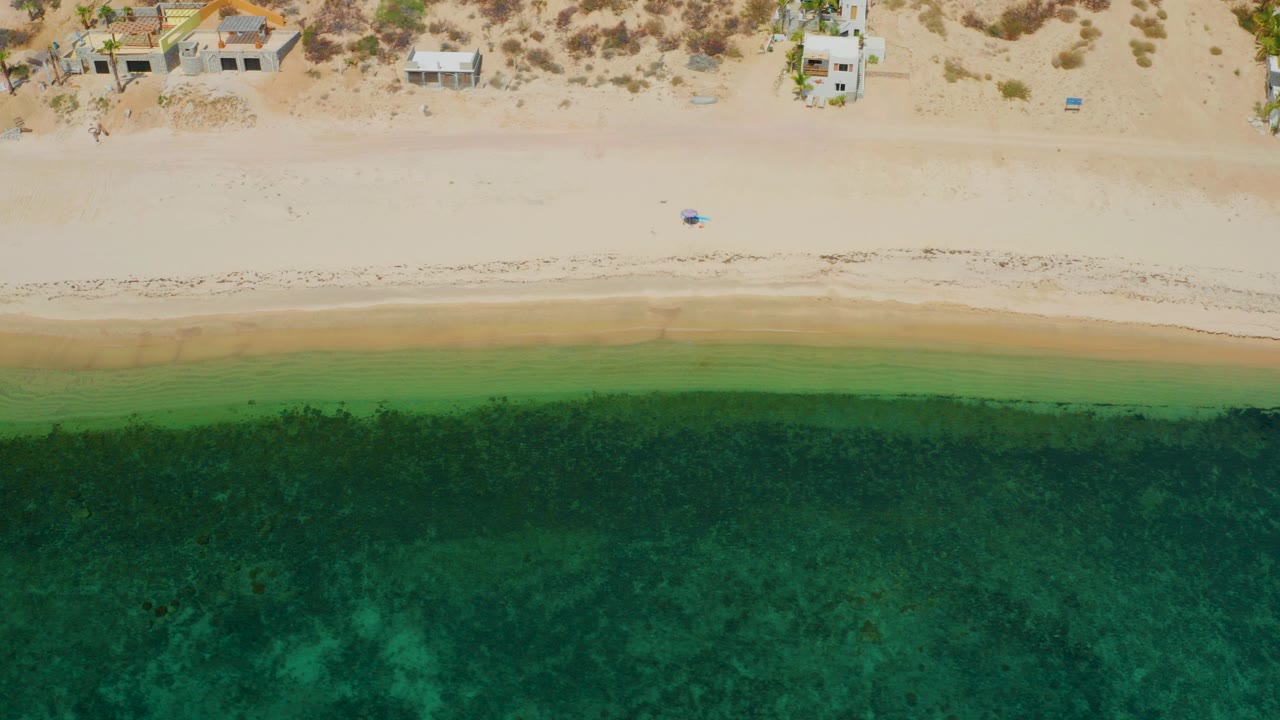 Overhead view in El Sargento La Ventana, Mexico showing dark green water below a pale beach and small shoreline homes at the top, with no visible people.