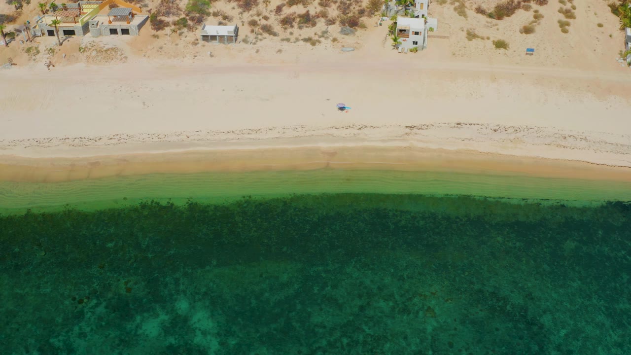 Aerial frame over El Sargento La Ventana, Mexico with a broad pale sand strip, green shallows, darker sea, and a few buildings near dry vegetation, with no visible people.
