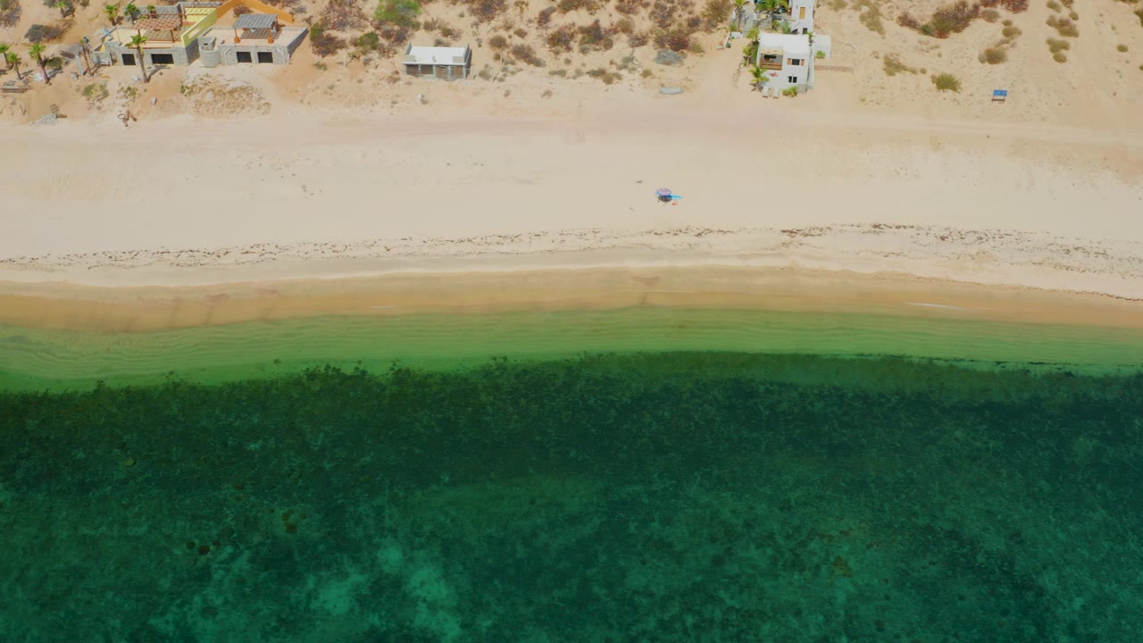 Top-down coastal view in El Sargento La Ventana, Mexico showing open beach between small homes and layered green water, with no visible people.