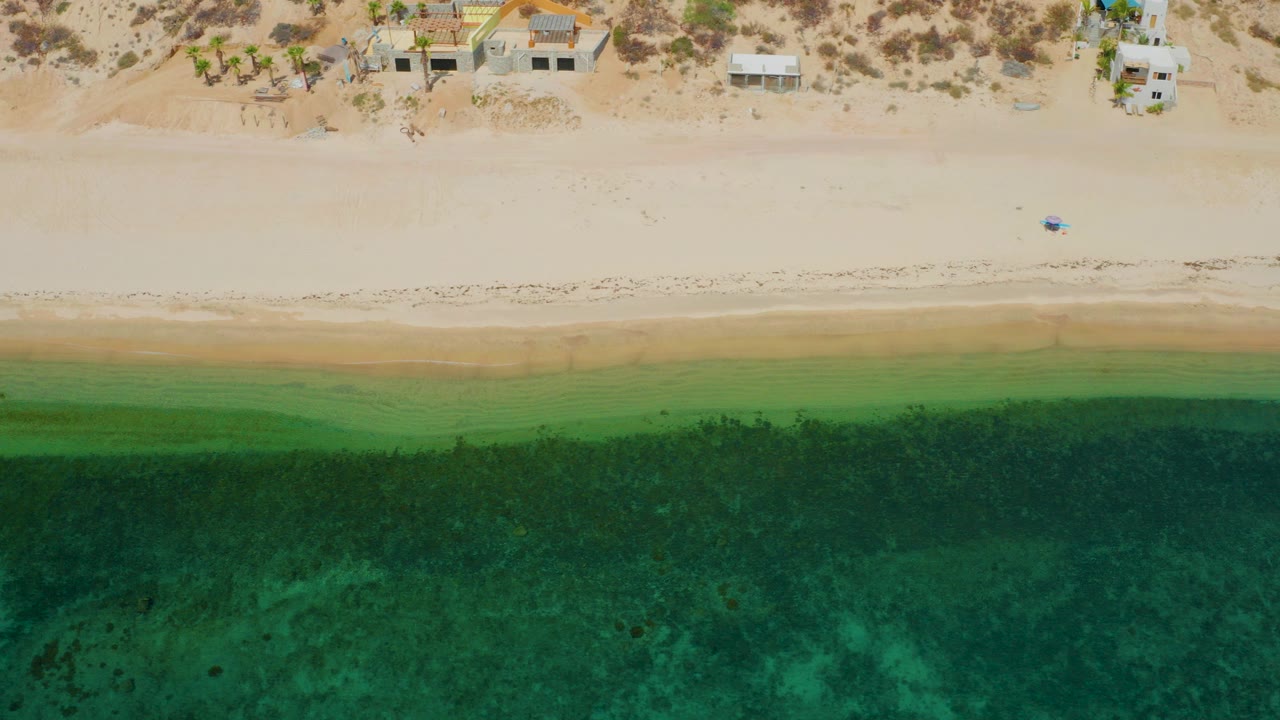 Top-down view of El Sargento La Ventana shoreline with tan sand, green shallow water, darker algae patches, small homes near the beach edge, and a tiny person on the sand.
