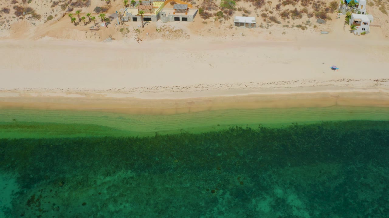 Overhead view in El Sargento La Ventana with bright sand, subtle dark and light shoreline bands, green water below, and small coastal structures along the top edge.