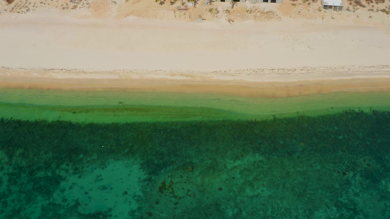 Aerial view over El Sargento La Ventana with the beach edge running across the upper frame and darker underwater patches spread through the green sea, no visible people.
