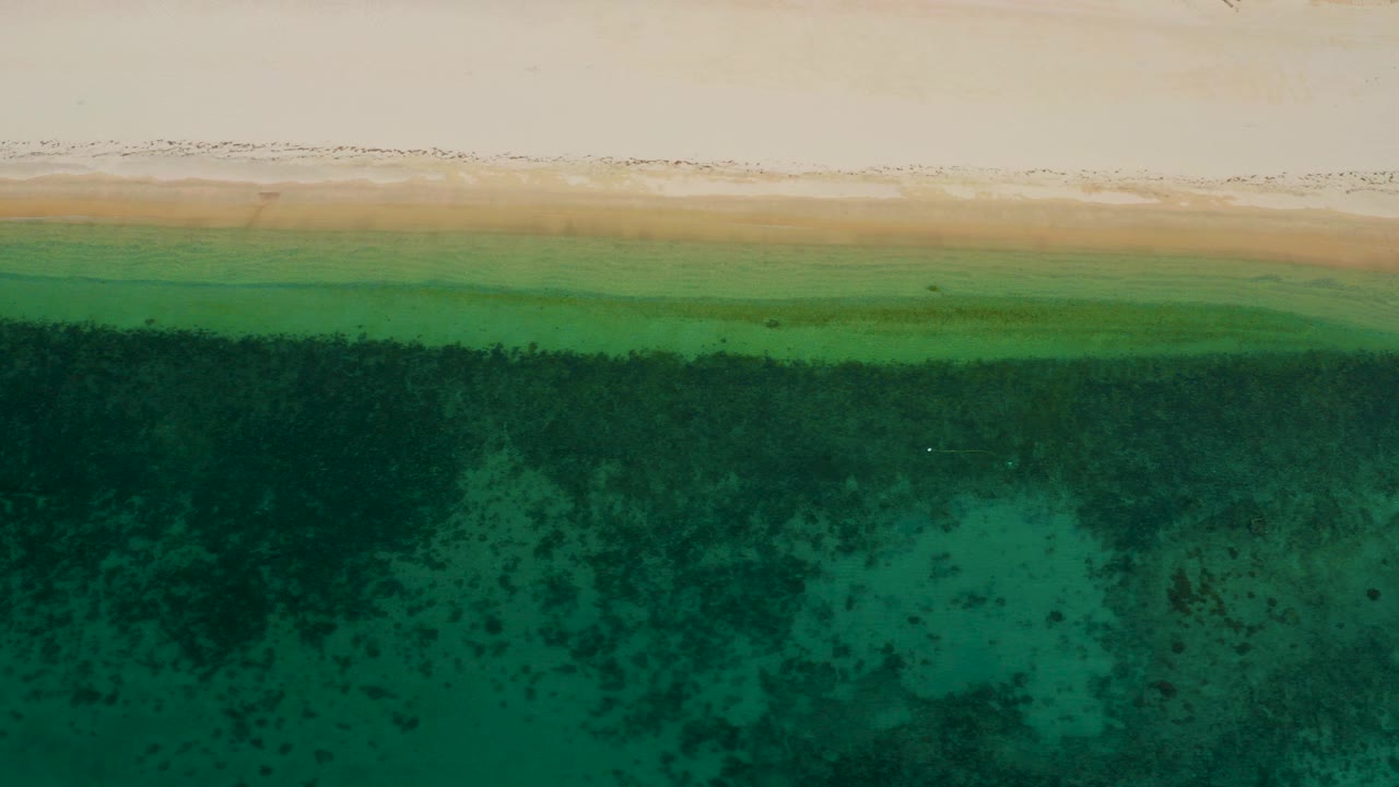 Overhead frame from El Sargento La Ventana, Mexico showing pale beach sand, a thin line of surf, turquoise-green shallows, darker underwater patches, and no visible people.