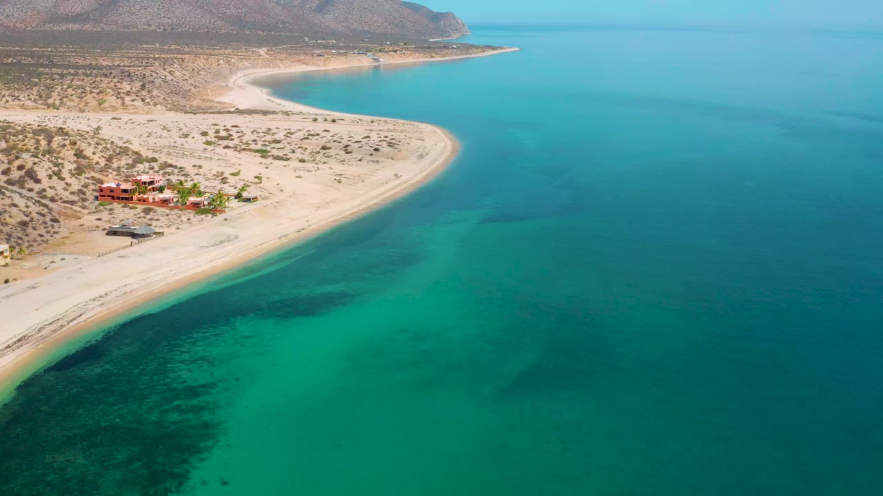 At 0:00, an aerial view over El Sargento La Ventana, Mexico shows a sandy curve of beach beside turquoise water, desert shrubs, distant mountains, red coastal buildings, and no visible people.