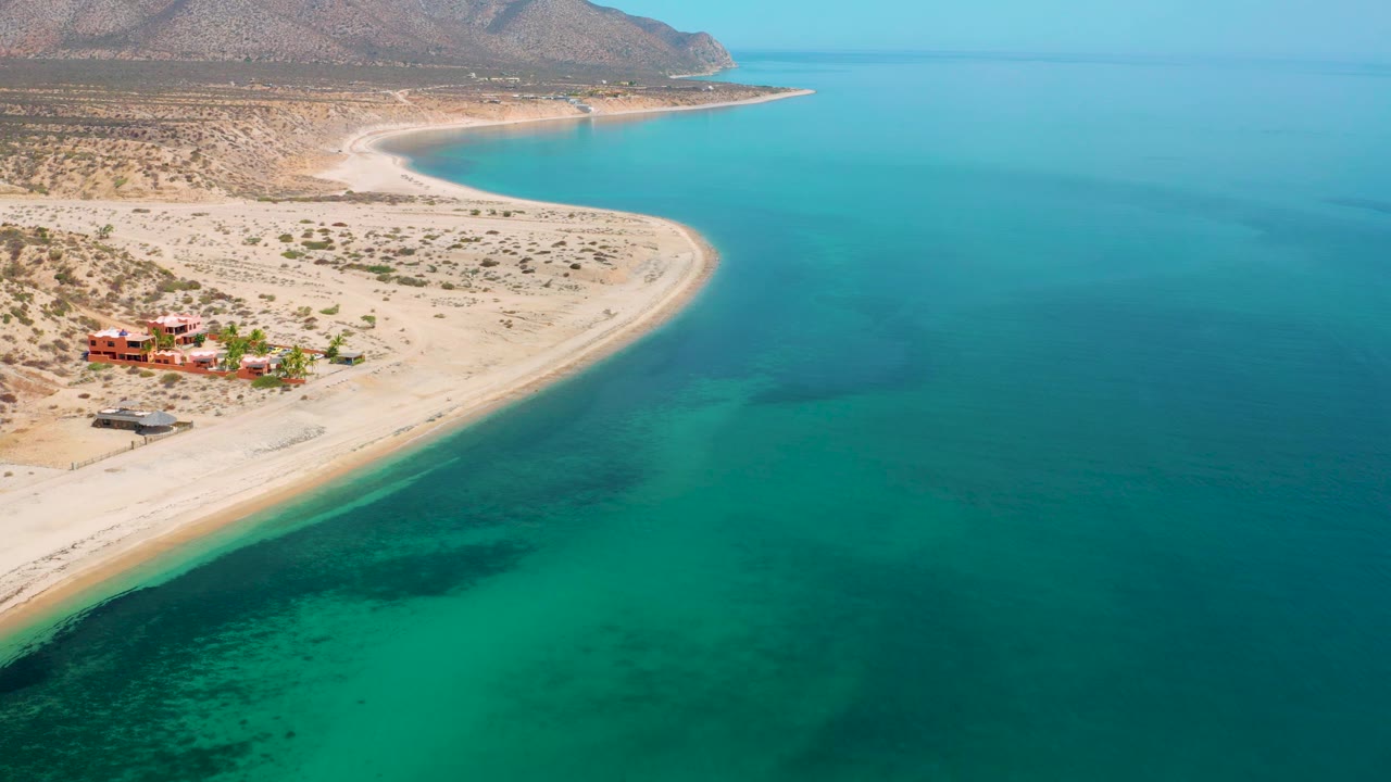 Wide aerial view in El Sargento La Ventana, Mexico, showing the shoreline sweeping from the lower left toward distant headland, clear turquoise sea, sparse desert vegetation, and no visible people.