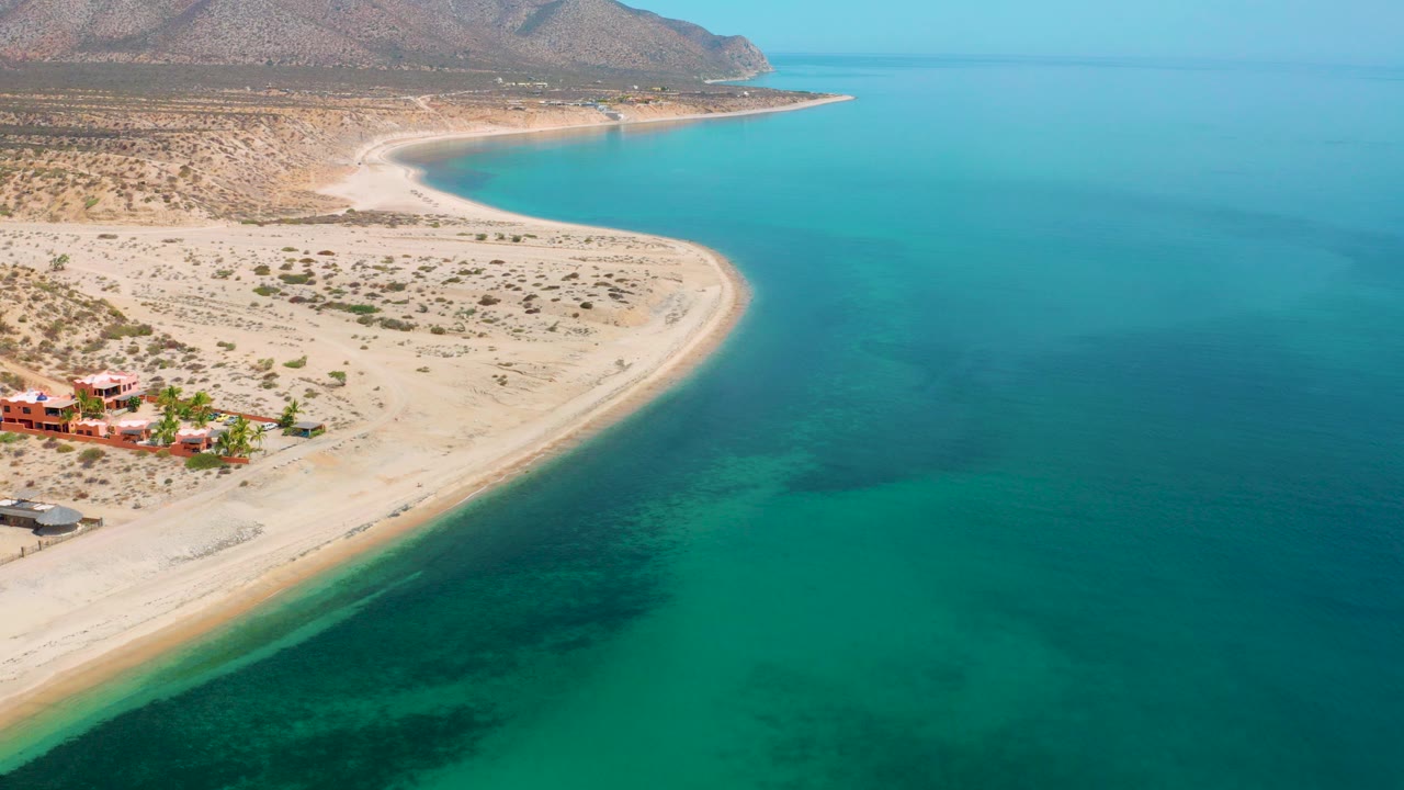 Opening aerial frame over El Sargento La Ventana, Mexico, with red-roofed buildings at lower left, a pale curved beach, turquoise water to the right, distant hills, and no visible people.