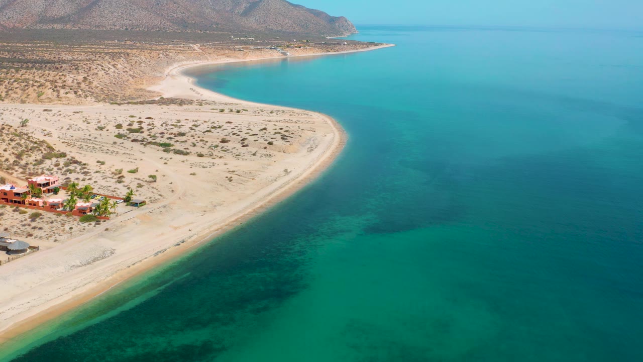Wide aerial frame over El Sargento La Ventana, Mexico, with the sandy point reaching into blue water, darker underwater areas near shore, arid land behind it, a few buildings at lower left, and no visible people.