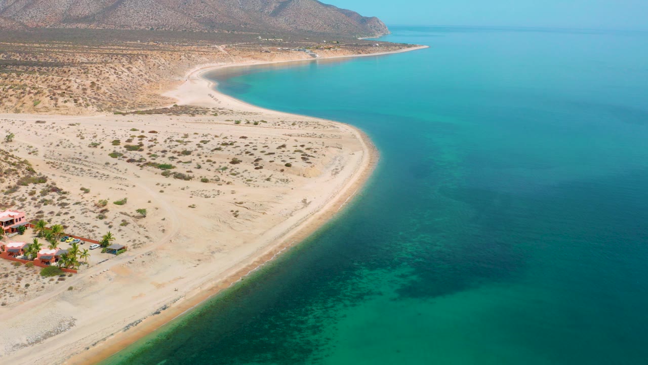 Aerial frame over El Sargento La Ventana, Mexico, showing a pale sand curve, blue-green water, dry scrubland, distant mountains, and no visible people.