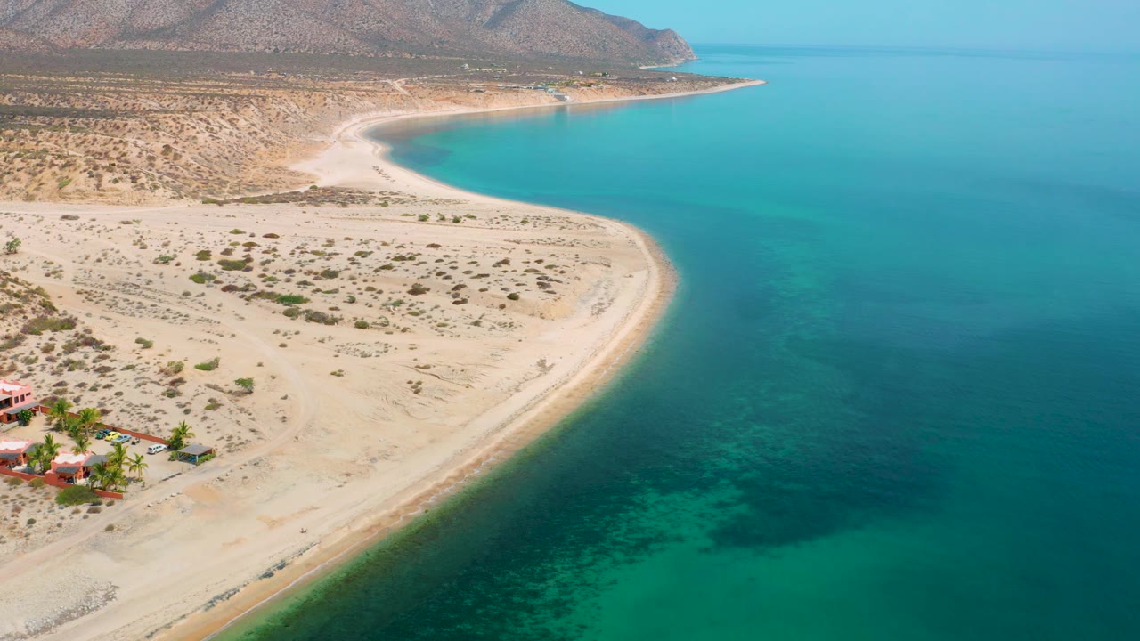 Wide aerial frame from El Sargento La Ventana, Mexico, showing blue water wrapping a pale sandy shoreline, sparse vegetation inland, low mountains, and no visible people.