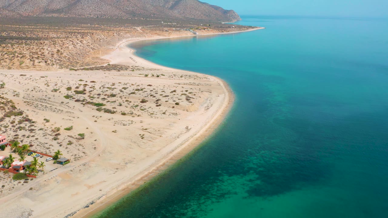 High coastal view with a beach curve, turquoise shallows, scrubland, and hills