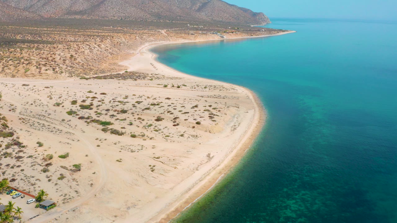Aerial view over El Sargento La Ventana, Mexico, showing a curved golden sand shoreline, turquoise green water, desert scrub, rocky mountains, and no visible people.