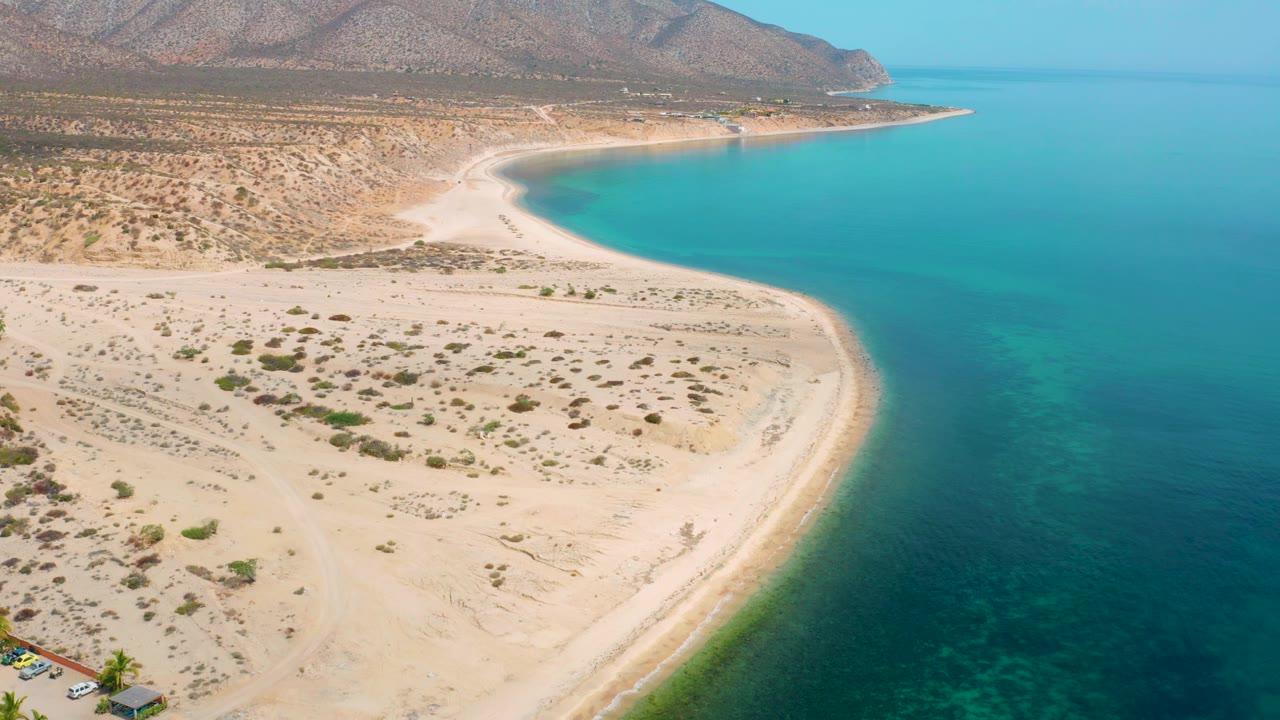 Aerial frame over El Sargento La Ventana, Mexico, with the sandy point more centered, turquoise sea to the right, desert scrub across the beach, mountains in back, and no visible people.