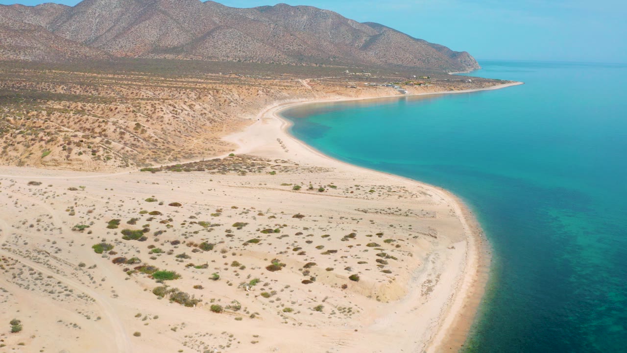 Aerial view of sandy beach curving beside blue water and desert mountains.