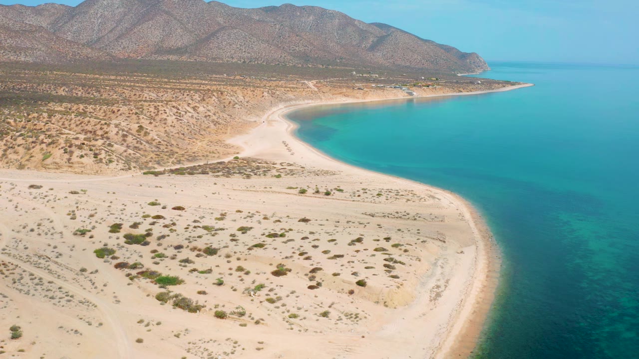 Aerial view in El Sargento La Ventana, Mexico, with the shoreline arc held near center, sandy desert foreground, clear blue-green water along the right edge, mountain ridges beyond, and no visible people.