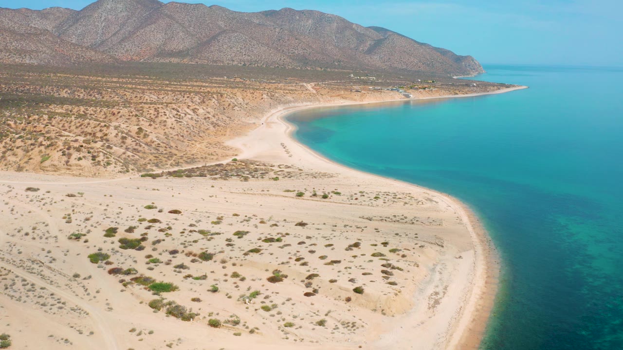 El Sargento La Ventana, Mexico, appears from above with more open water on the right, a pale beach wrapping around the bay, scattered shrubs on the sand, rugged mountains behind, and no visible people.